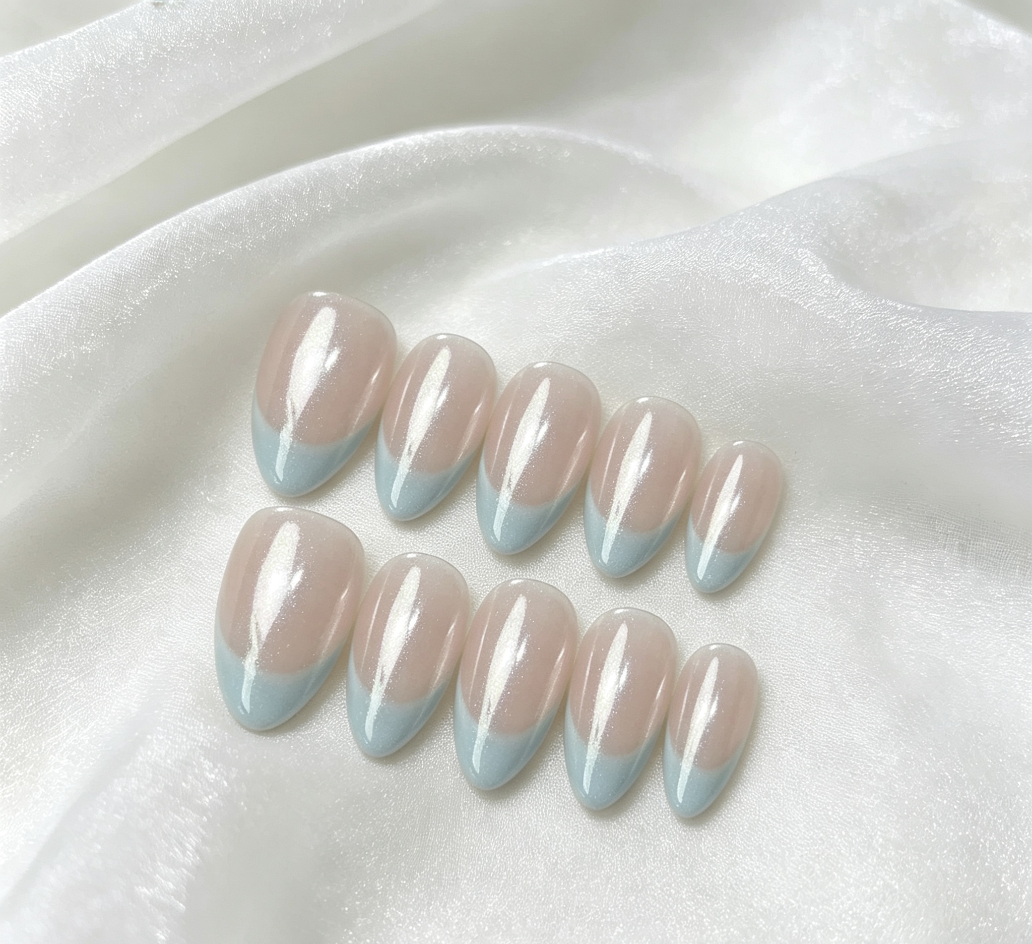 Two rows of almond-shaped press-on nails with nude bases and pale blue tips on white fabric.